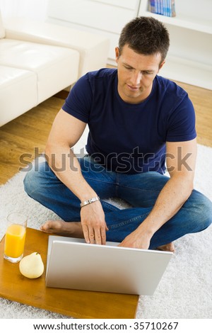 Casual young man working at home on his laptop computer, sitting on floor, looking at screen. High angle view.