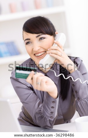Smiling young women shopping online at home, holding credit card in hand and talking on phone.