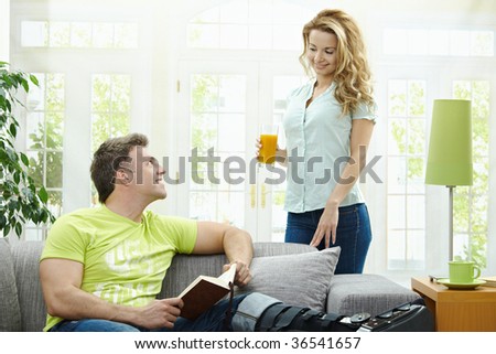 Man rasting his broken leg in cast on sofa at home, reading book. Woman bringing him orange juice.