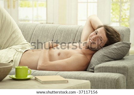 Casual bare chested young man resting on couch at home, smiling.