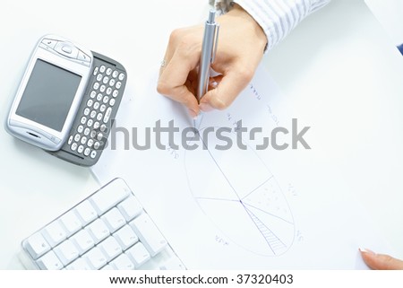Female hand holding pen, drawing pie chart, beside desktop computer keyboard and mobile phone.