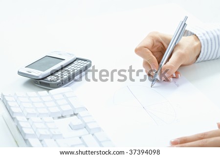 Female hand holding pen, writing on paper, beside desktop computer keyboard and mobile phone.