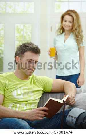 Man rasting his broken leg in cast on sofa at home, reading book. Woman bringing him orange juice.