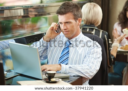 Businessman sitting at table in cafe using laptop computer, talking on mobile.