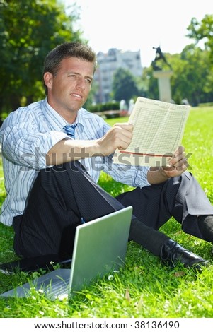Relaxed businessman sitting in grass beside laptop computer, reading newspaper.
