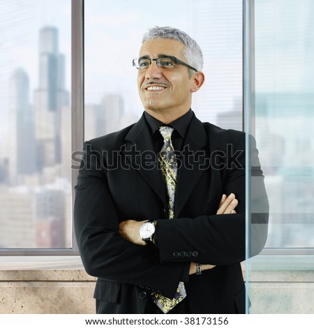 Confident businessman standing with arms crossed in front of office windows, smiling.
