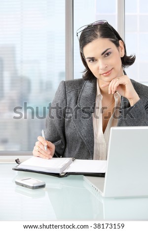 Businesswoman sitting at desk in front of offive windows, thinking over laptop computer and personal organizer.