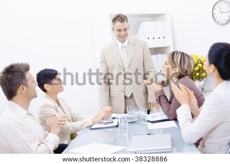 Group of businesspeople sitting at table in meeting room, clapping and looking at smiling businessman.