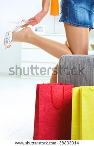 Woman taking off high heel shoe at home. Colorful shopping bags in front.