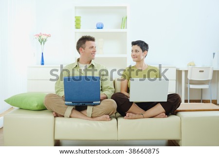 Happy young couple sitting on couch at home using laptop computer, smiling.