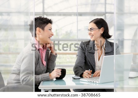 Happy young businesswomen working together at desk in modern office, looking at each other, laughing.