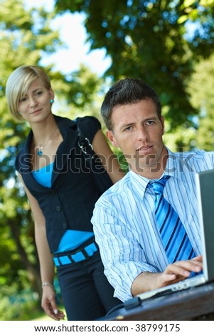 Casual businessman using laptopn in park summertime, young woman watching from behind.