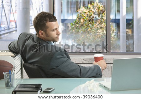 Relaxed businessman sitting in front of windows,  holding coffee cup looking back to laptop on office desk.