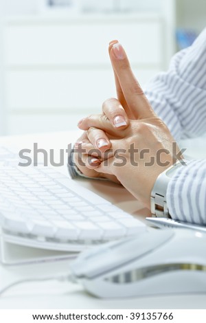 Closeup of female hands with fingers crossed at desk beside computer keyboard.