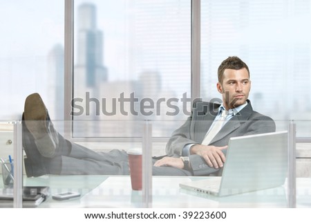 Casual businessman sitting at desk in front of office windows, thinking.