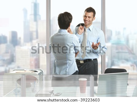 Two businesspeople standing at desk in downtown office building, talking and smiling.