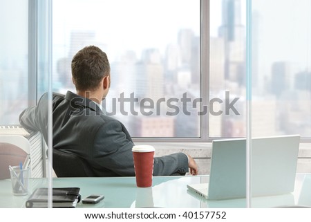 Relaxed businessman sitting at desk in office, looking out the windows to downtown skyscrapers.