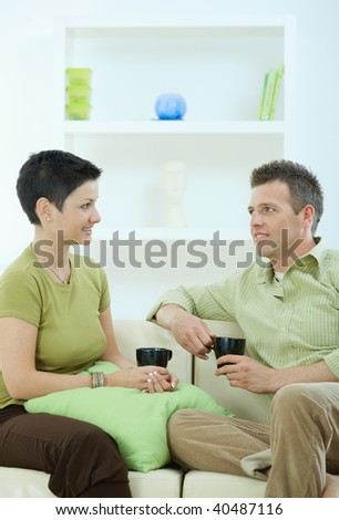 Young couple drinking coffee at home, sitting on couch, looking at each other, smiling.