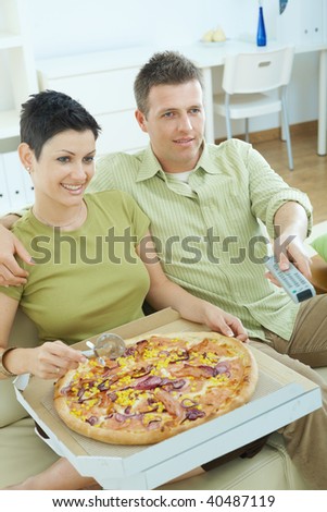 Happy couple sitting on sofa eating pizza and watching TV at home, smiling.