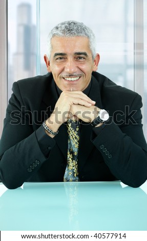 Portrait of casual businessman, sitting at desk in front of office windows, leaning on hands, smiling.