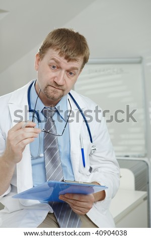Medical office - troubled looking male doctor sitting on desk, holding clipboard, thinking.