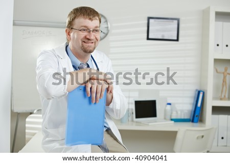 Medical office - happy male doctor sitting on desk,holding file, looking away, smiling.