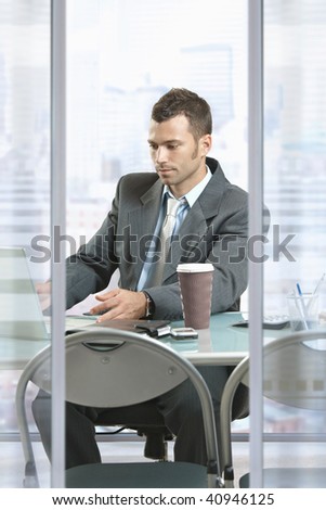 Profile portrait of businessman sitting at desk in front of office windows, using laptop computer.