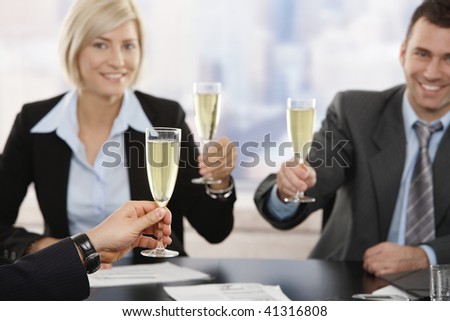 Happy young business people sitting around meeting table at office raising toast with champagne, smiling. Focus placed on hand in front.