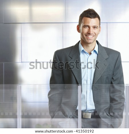 Confident businessman standing in modern office with glass and metal walls, smiling.