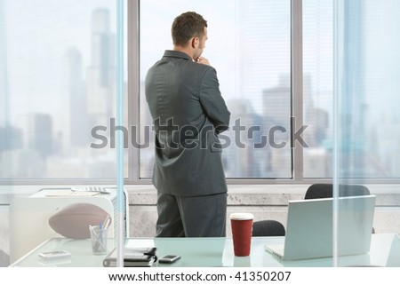 Businessman thinking in office, looking out of windows to downtown vista.