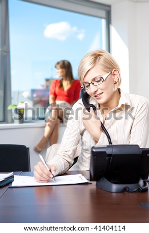 Young businesswoman sitting at desk in office, talking on landline phone, writing notes to personal organizer.