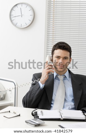 Businessman sitting at desk in office, talking on landline phone, smiling.