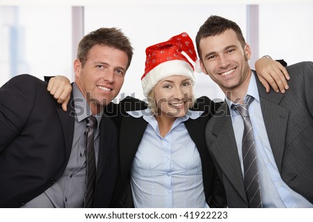 Happy young businesswoman wearing Santa Claus hat at office hugging friends, looking at camera, smiling.