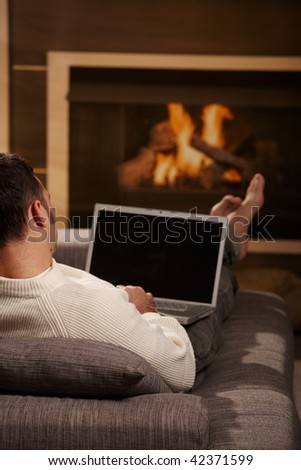 Man sitting on sofa at home in front of fireplace and using laptop computer, rear view.