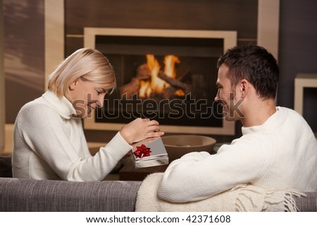 Young romantic couple sitting on couch in front of fireplace at home, man giving gift, side view.