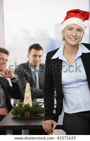 Happy young businesswoman wearing Santa Claus hat at office, looking at camera, smiling. Businessmen in background.
