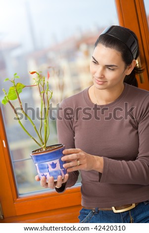 Attractive young woman standing at window, holding green potted plant, smiling.