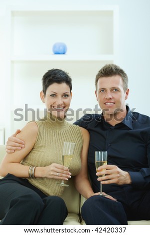 Love couple clinking champagne glasses at home on sofa. Smiling and looking at camera.