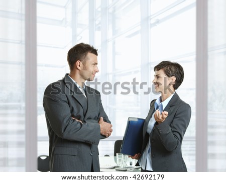 Businessman and businesswoman leaving meeting room, talking at the door, smiling.