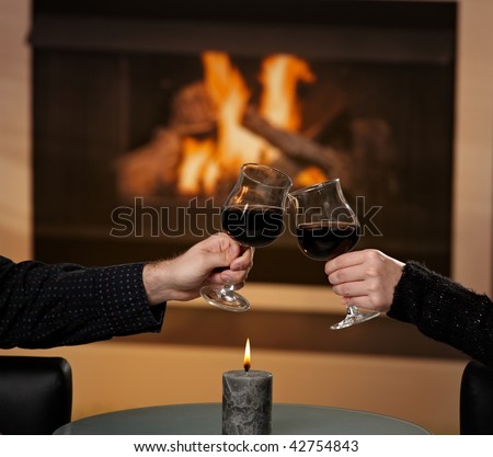 Hands holding glass of red wine, clinking in front of fireplace.