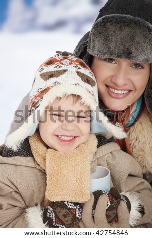 Portrait of happy mother and child holding cup of hot tea in snow on a cold winter day laughing, smiling.