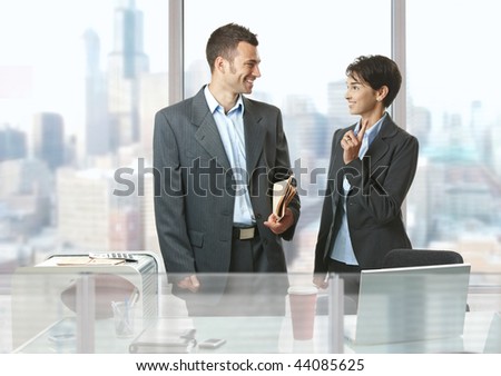 Two businesspeople standing at desk in front of windows in office, talking and smiling.