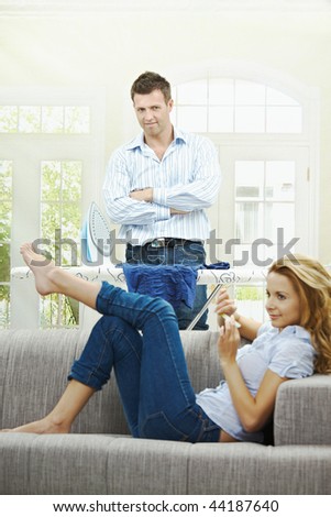 Relaxed young woman sitting on couch filing her nails, man watching with hands crossed behind ironing board. Focus on man.