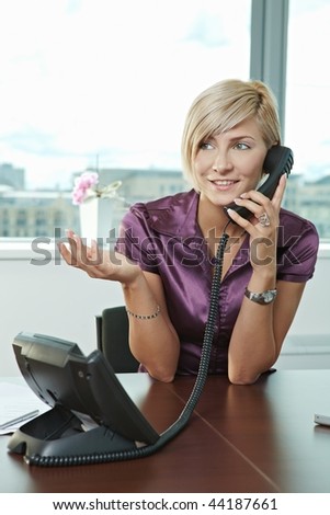 Happy young businesswoman sitting at table in office meeting room, talking on phone, smiling.