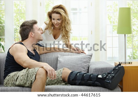 Love couple at home. Man resting his broken leg on sofa, his girlfriend embracing him from behind.