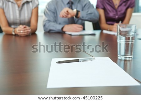 Focus on paper and pen on table. In the background panel of business people sitting conducting job interview.