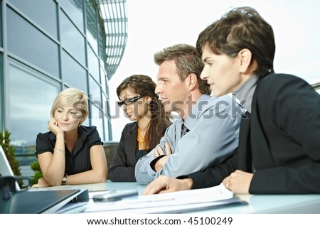 Group of young business people sitting in a row at table on office terrace outdoor, talking and working on laptop computer.