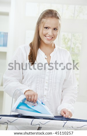 Happy young woman ironing on ironing board at home, smiling, looking at camera.