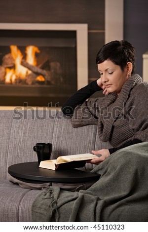 Woman sitting on sofa at home on a cold winter day, reading book.