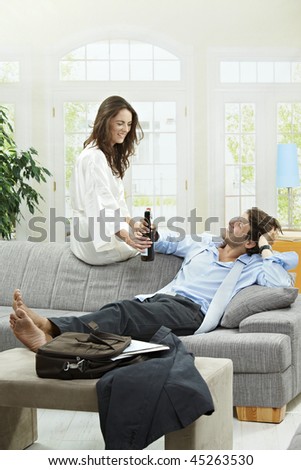 Tired businessman resting on couch at home after long day of work. Woman giving him bottle of beer.